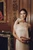 Model in a white satin dress holding the closed, sculptural walnut wood Petite Bird handbag, photographed in an elegant wood-paneled interior. A delicate wood-and-metal flower bud brooch is pinned to her dress.