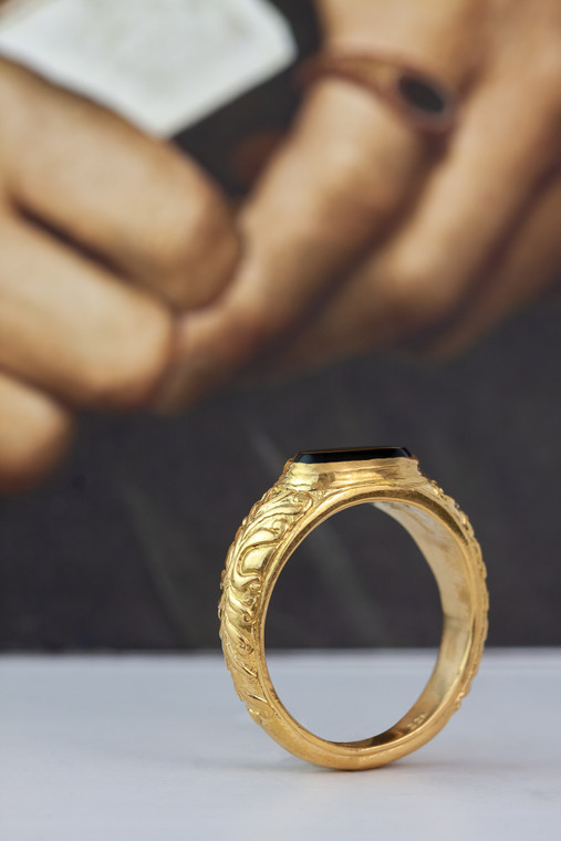 Gold ring with black onyx stone standing up in front of a backdrop of Sir Thomas More's fingers.