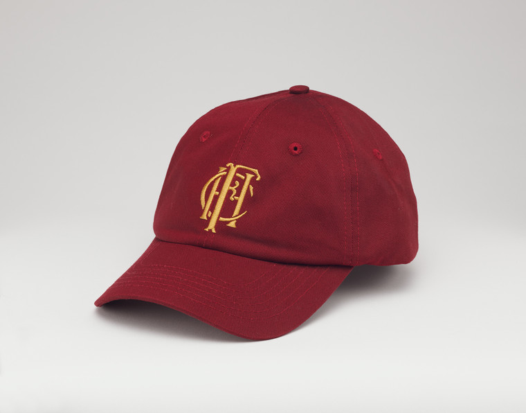 Burgundy baseball hat with the embroidered gold Frick Collection logo on the front sitting on a white surface.