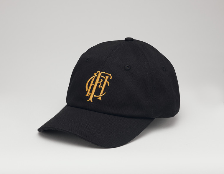 Black baseball hat with the embroidered gold Frick Collection logo on the front sitting on a white surface.
