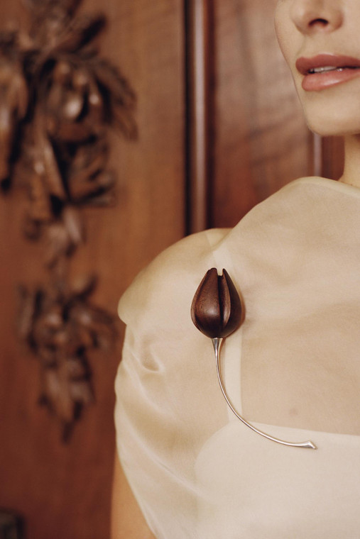 Model wearing a cream blouse with a carved wood (walnut) flower bud brooch pinned at the shoulder, featuring a slender curved silver stem.