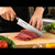 Aritsugu Japanese Gyuto chef knife slicing fresh beef on a wooden cutting board in a professional kitchen, handcrafted Japanese kitchen knife in use.