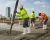 Construction workers wearing white rubber boots while pouring and finishing concrete at ground level with high-rise buildings in the background.