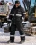 Worker wearing a black insulated reflective winter coverall, safety glasses, gloves, and work boots at a snowy industrial jobsite with heavy equipment.