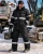 Worker wearing a black insulated reflective winter coverall, safety glasses, gloves, and work boots at a snowy industrial jobsite with heavy equipment.