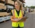 Blonde worker wearing an ANSI Class 2 reflective safety vest while inspecting materials in a lumber stock yard
