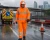 Roadwork crew member wearing a hi-vis orange waterproof rainsuit with reflective tape while working in rainy highway maintenance conditions.
