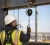 Construction worker wearing a fall protection harness, hard hat, and hi-vis vest while using a self-retracting lifeline attached to a steel anchor point.