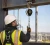 Construction worker wearing a fall protection harness, hard hat, and hi-vis vest while using a self-retracting lifeline attached to a steel anchor point.