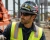 Construction worker wearing a patriotic eagle graphic hard hat, safety sunglasses, and hi-vis vest at an active industrial jobsite.