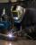 Industrial welder wearing a shark graphic welding helmet, welding gloves, and protective workwear while welding metal in a workshop