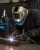 Industrial welder wearing a shark graphic welding helmet, welding gloves, and protective workwear while welding metal in a workshop