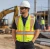 Construction worker wearing a hi-vis surveyor safety vest, white hard hat, safety sunglasses, and work pants at an active outdoor jobsite
