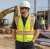Construction worker wearing a hi-vis surveyor safety vest, white hard hat, safety sunglasses, and work pants at an active outdoor jobsite