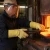 Metal workshop worker wearing yellow ThermoBest heat-resistant gloves, face shield, and protective workwear while handling hot metal near a furnace