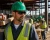 Construction worker wearing a green hard hat, safety sunglasses, hi-vis vest, and work gloves while carrying lumber