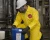 Industrial worker wearing a yellow Tychem chemical protective suit, respirator, hard hat, safety glasses, and gloves while handling a chemical container in a plant