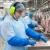 Food processing worker wearing safety glasses, earmuffs, beard cover, disposable gown, sleeve covers, and gloves while handling raw meat.