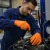 Worker wearing orange nitrile disposable gloves while performing industrial maintenance on hydraulic equipment.
