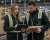 Male and female warehouse workers wearing black hi-vis safety vests while reviewing inventory.