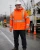 Older male utility worker wearing a high-visibility orange arc flash rain jacket with reflective striping on a wet roadside worksite.