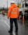 Back view of an older male utility worker wearing a high-visibility orange arc flash rain jacket with reflective striping and hood on a rainy roadwork site.