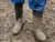 Worker wearing black rubber work boots covered in mud while standing on a wet construction or agricultural jobsite.