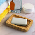 A bamboo soap holder in a wooden tray with a white soap bar, surrounded by towels and a glass container.