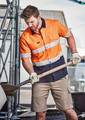 A short sleeve work shirt in orange and navy with reflective tape, worn by a man using a shovel, with a construction setting in the background.