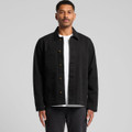 A man wearing a black canvas chore jacket with button closure and multiple pockets. The background is plain grey.