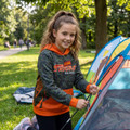 A girl in a camouflage and orange hoodie stands beside a tent in a park, smiling at the camera.