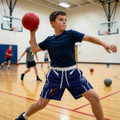 A boy wearing blue sports shorts and a black t-shirt throws a red ball in a gym setting. The image features other children in the background.