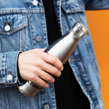 A drink bottle in stainless steel with a sleek design, held by a person in a denim jacket. It features a logo.