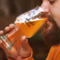 A beer glass with a logo, featuring a curved design, holds a golden beverage near a person's face.