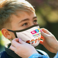 A reusable 3-ply face mask featuring colourful graphics, being fitted onto a child's face outdoors.