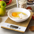 A bamboo kitchen scale with a white bowl containing flour, sugar, and an egg, surrounded by fresh ingredients.