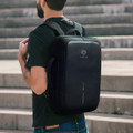 A black anti-theft backpack with a sleek design, featuring a logo and a top handle, worn on a person’s back.