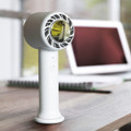 A white personal cooling fan sits on a wooden table, with a blurred electronic device in the background.