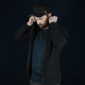 A man in a black Swiss Peak Urban Hoodie adjusts the hood, wearing a black cap. The background is dark.