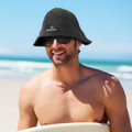 A man wearing a black bucket hat and sunglasses, sitting on a beach with ocean waves in the background.