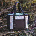 A cooler bag in grey and black with sturdy handles and a front pocket, hanging from a tree branch.