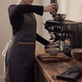 A dark grey canvas apron with an adjustable strap, worn by a person near a coffee machine, alongside wooden utensils and bowls.