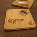 A square bamboo coaster featuring a bottle opener and a logo, set on a wooden surface with a drink.