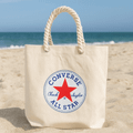 A cream-coloured tote bag with a round logo featuring a red star and branding, set against a beach background.