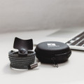 A charging cable in black and white fabric with a snap closure, packaged in a circular black case with a logo.