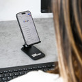 A black folding desktop mobile phone stand holding a smartphone, positioned on a desk near a computer keyboard.