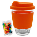 A clear glass coffee cup with an orange silicone sleeve and lid, accompanied by a pack of colourful jelly beans.