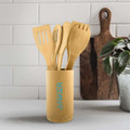 A bamboo cutlery set consisting of various utensils, displayed in a cylindrical holder, on a countertop with a plant and cutting board.