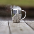 A silver coffee mug with a reflective surface and steam rising, placed on a wooden table. It features a logo.