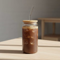 A glass tumbler with a bamboo lid and straw, featuring dark beverage and ice cubes, set on a wooden table.